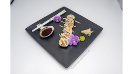 A sushi roll arranged on a black square plate with 2 purple flowers, wasabi, pickled ginger, a spoon with soy sauce and chopsticks.