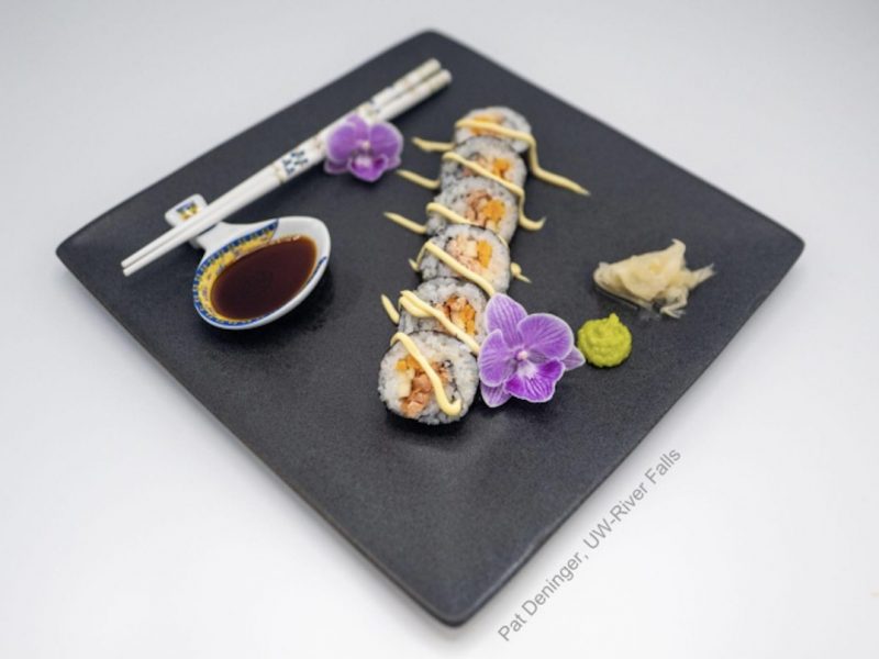 A sushi roll arranged on a black square plate with 2 purple flowers, wasabi, pickled ginger, a spoon with soy sauce and chopsticks.