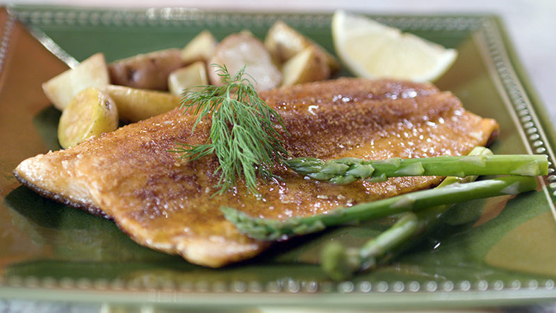 This is a photograph of seared fish accented by asparagus, lemon, potatoes and dill on a green plate.