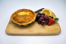 A small pie decorated with a fish-shaped piece of crust sits atop of a cutting board next to grapes, a strawberry and a lemon wedge.